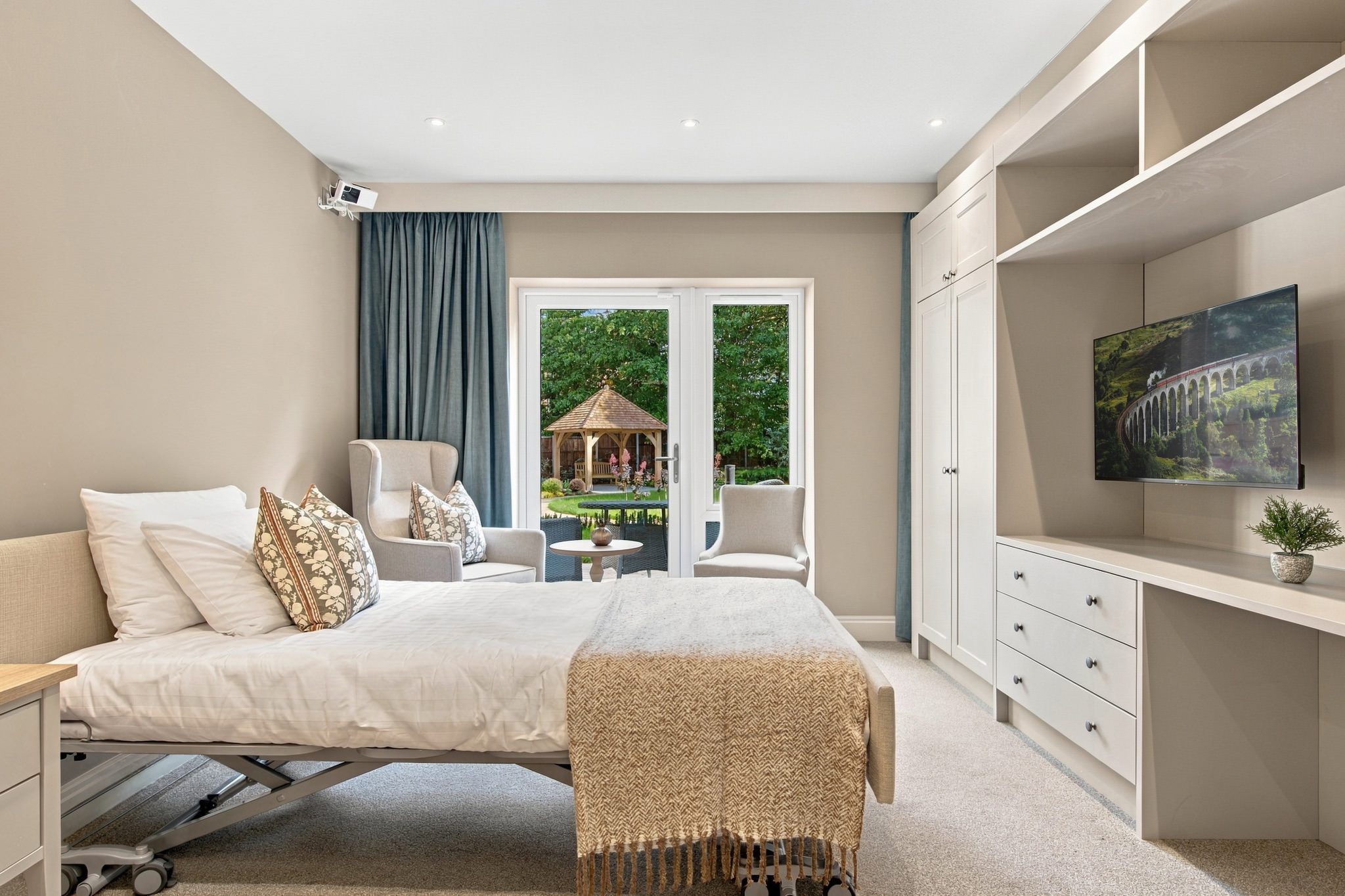Resident bedroom at Kings Hill with a comfortable bed, armchairs and patio doors opening onto a garden with a gazebo.
